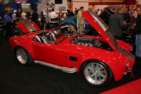 1998 Ford Crown Victoria - A red convertible sports car with its hood open is displayed indoors with other vehicles and people.