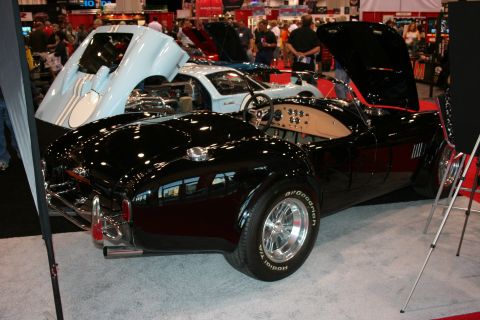1998 Ford Crown Victoria - A black AC Cobra is displayed with its hood open, revealing its tan interior and engine bay, alongside another car with its hood up.