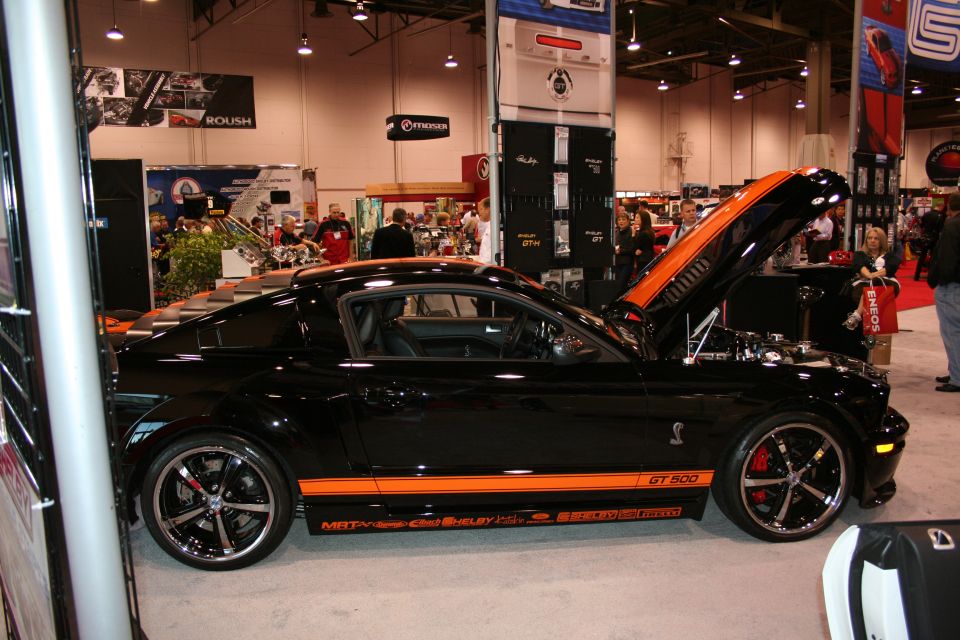 Black Ford Mustang Shelby GT500 with orange stripes, hood open, showing engine. Text on side reads 'GT 500' and 'Shelby'