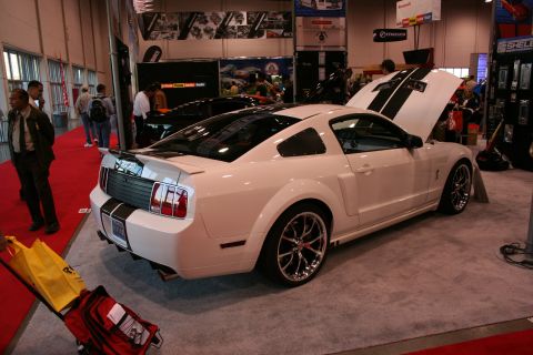 1998 Ford Crown Victoria - A white Ford Mustang with black racing stripes is displayed with its hood open at an auto show.