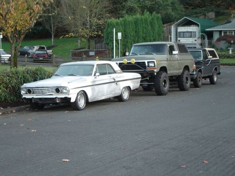 1989 Ford Bronco - A line of vehicles, including a vintage car and several lifted trucks, are parked on a residential street.