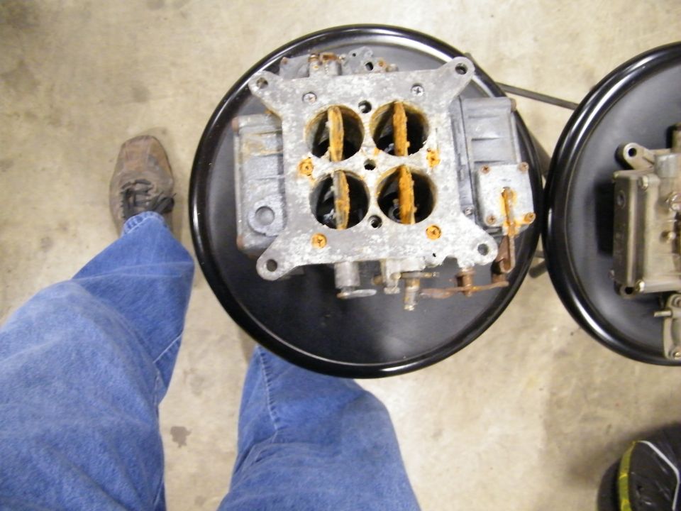 Two carburetors on black circular bases, one in focus, with a person's legs and shoes visible