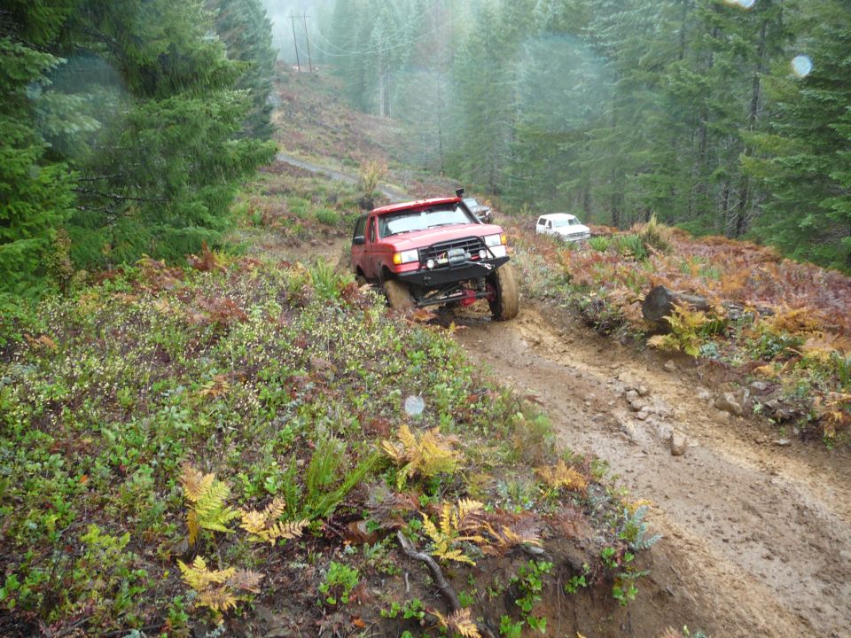 1991 Ford Bronco navigating a steep, muddy off-road trail with another white SUV behind it