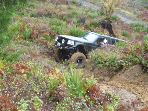 1991 Ford Bronco - A black SUV with large off-road tires is driving up a steep, muddy hill covered in vegetation.