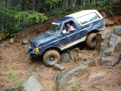 1991 Ford Bronco - A blue Ford Bronco with a white top is shown off-roading on a challenging rocky and muddy terrain.