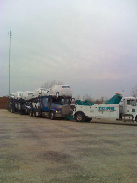 1992 Ford Bronco - A car carrier truck transports multiple vehicles, with a tow truck positioned nearby under an overcast sky.