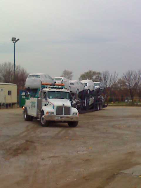 1992 Ford Bronco - Car carrier truck transporting several vehicles across a lot under an overcast sky.