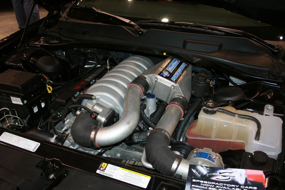 Engine bay with a supercharger and silver intake pipes. Black hoses connect to the supercharger. A yellow cap is visible on a fluid reservoir
