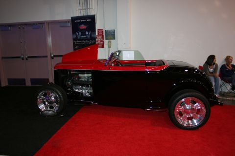 1998 Ford Crown Victoria - A two-tone hot rod roadster is displayed on a red carpet, its exposed engine and custom wheels.