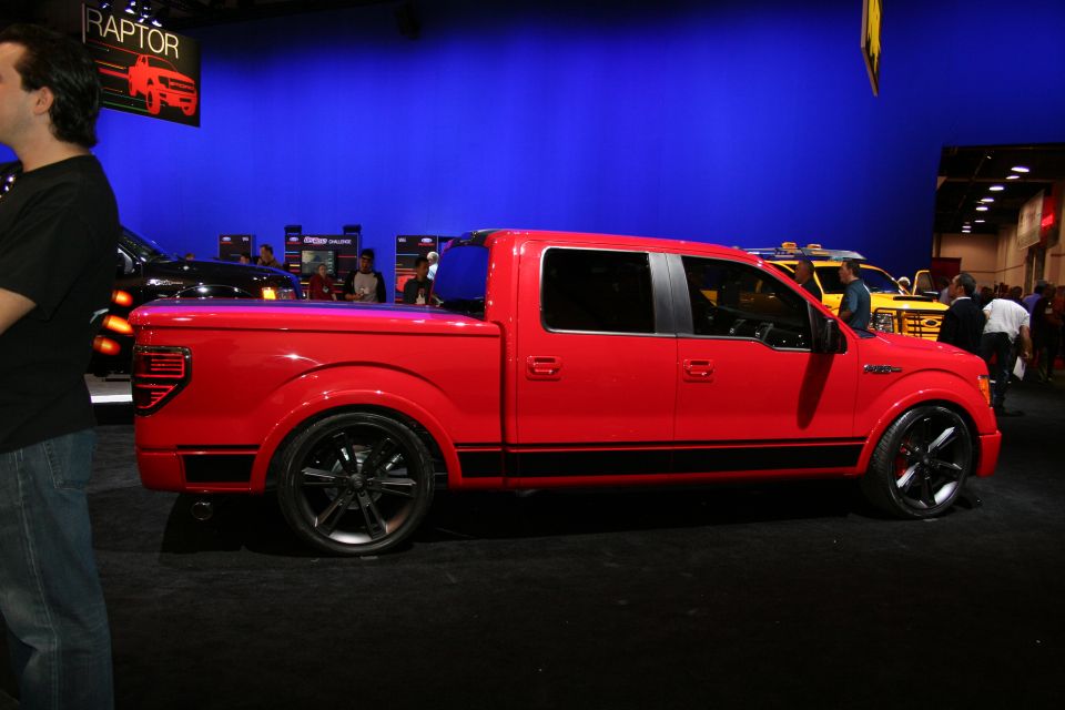 Red Ford F-150 pickup truck, side view, with black racing stripes and custom wheels. Another yellow truck is visible behind it