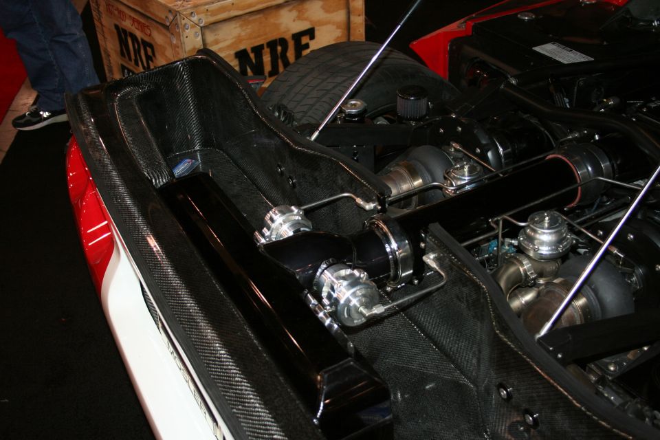 Engine bay with twin turbos, intercooler piping, and carbon fiber accents. A wooden crate with 'NRF' text is in the background