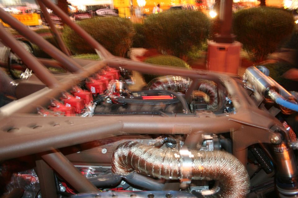 Engine bay with red ignition coils and metallic exhaust pipes, surrounded by a brown roll cage