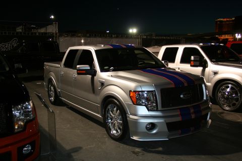 1998 Ford Crown Victoria - Modified silver Ford F-150 pickup truck with racing stripes is displayed at night next to other vehicles.
