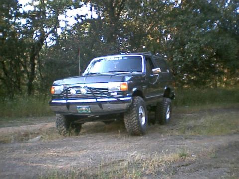1988 Ford Bronco - A lifted Ford Bronco sits on a dirt trail surrounded by trees.