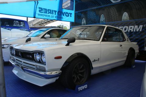 1998 Ford Crown Victoria - A white 1973 Nissan Skyline GT-R is displayed at an event with other vehicles and tire branding visible.