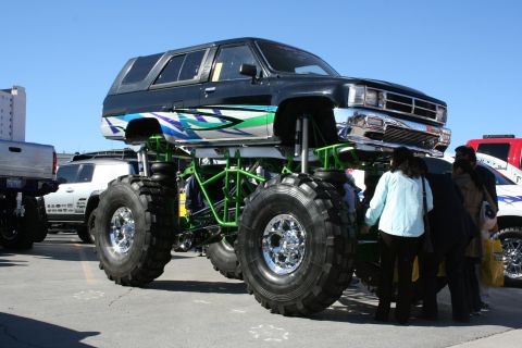 1998 Ford Crown Victoria - A custom monster truck with large tires and colorful graphics is displayed outdoors, with other vehicles and people nearby.