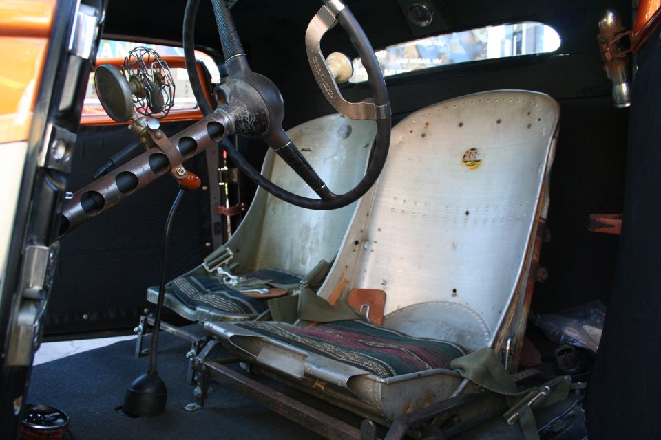 Interior view of a custom hot rod showing airplane seats, a vintage steering wheel, and a small fan mounted on the column