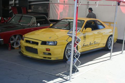 1998 Ford Crown Victoria - A yellow coupe with racing decals is parked next to a red and black car under a tent.