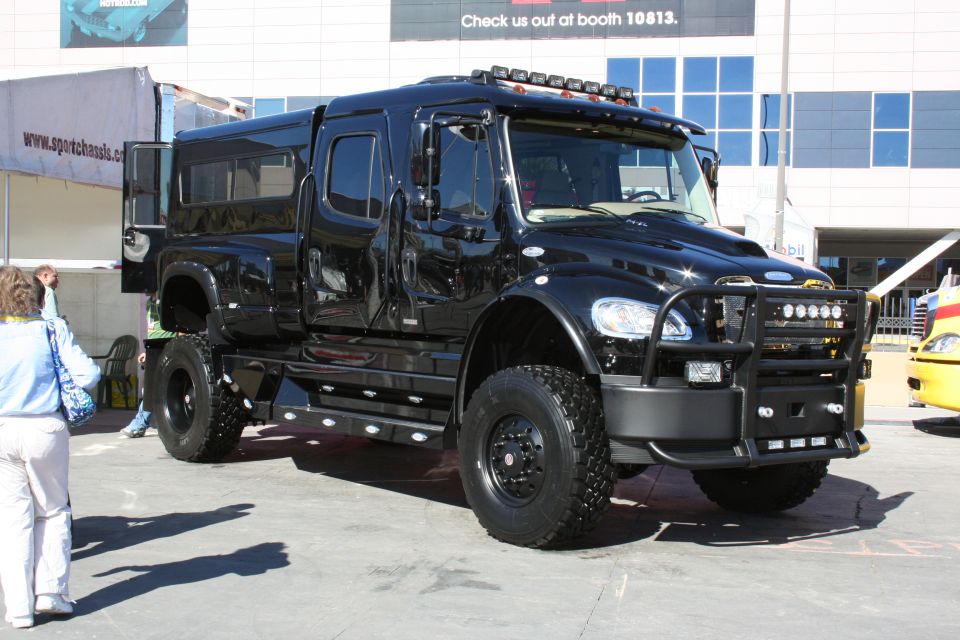 Black custom heavy-duty truck with crew cab, utility bed, and bull bar. Text on building reads 'Check us out at booth 10813.'