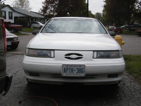 1993 Ford Taurus - A white sedan is parked in a driveway with other vehicles visible in the background.