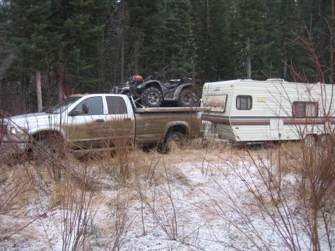 2005 Dodge 3500 - A muddy 2005 Dodge 3500 tows a trailer and ATV through a wooded area with patchy snow.