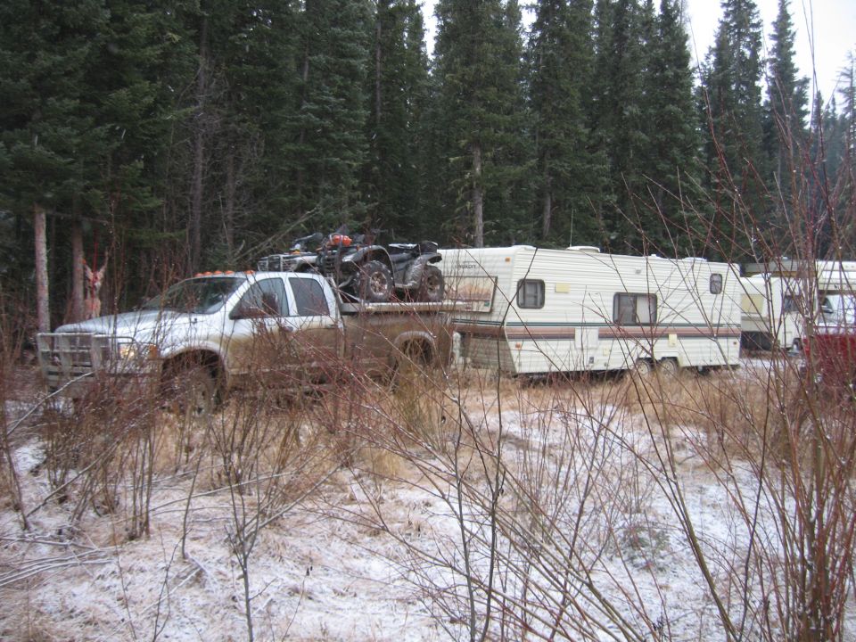 Silver pickup truck with a large grille guard, towing a white travel trailer. Two ATVs are loaded on the truck bed. Mud splatters on the truck