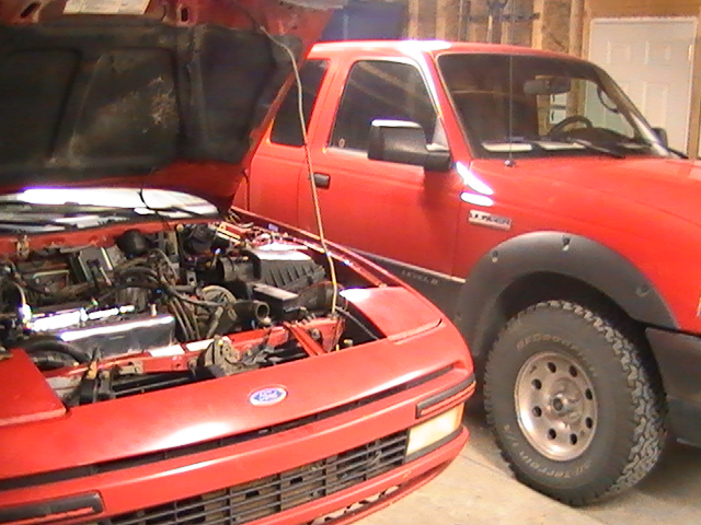 Red coupe with open hood and exposed engine next to a red Ford pickup in a garage.