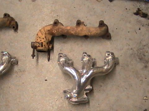 1990 Ford Probe - Two exhaust manifolds, one old and rusty, the other polished and new, lie on a concrete surface.