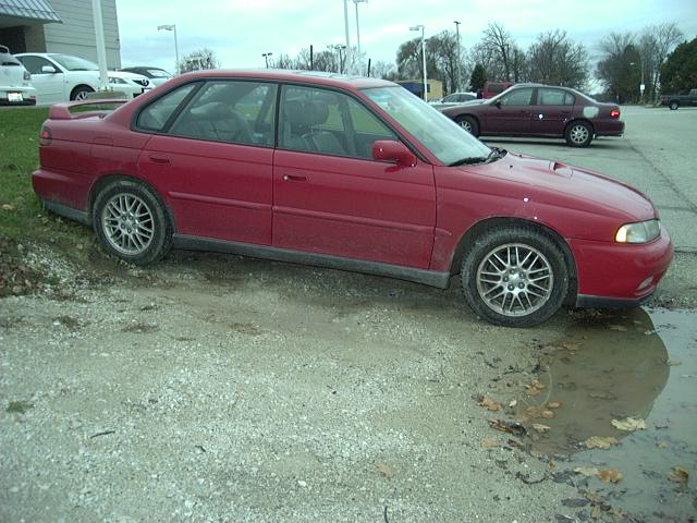 Side view of a dirty red Subaru Legacy sedan parked on a muddy gravel lot