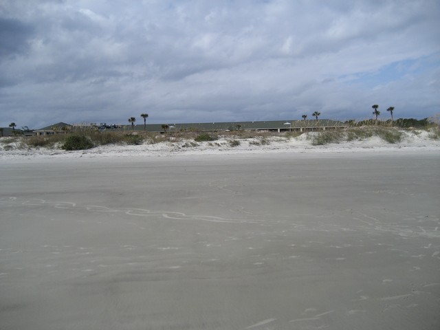 Beach with sand in foreground, dunes and buildings in background under cloudy sky