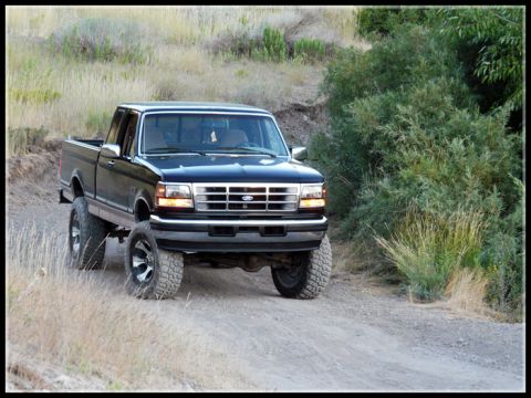 1996 Ford Bronco - This lifted black pickup truck navigates a dusty, off-road path surrounded by dry grass and brush.
