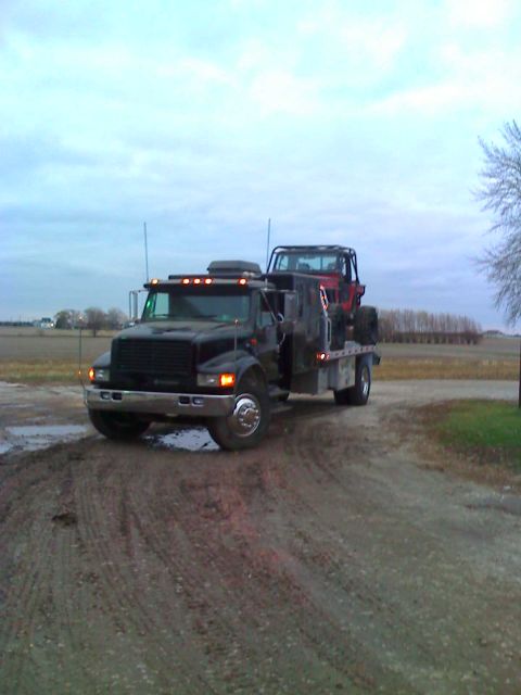 1992 Ford Bronco - A flatbed truck transports a red off-road vehicle across a muddy rural road under a cloudy sky.