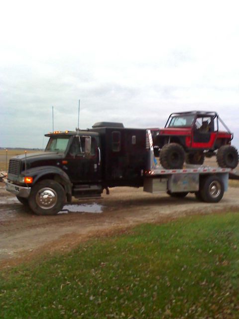 1992 Ford Bronco - A black flatbed truck transports a red off-road vehicle on its trailer.