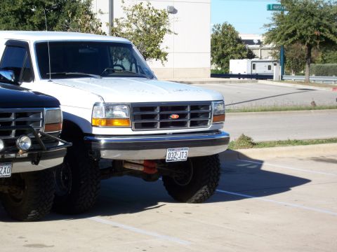 1996 Ford Bronco - A white 1996 Ford Bronco is parked next to another vehicle, showing its front grille and license plate.