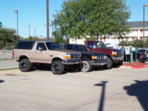 1996 Ford Bronco - Three lifted Ford trucks, including a 1996 Bronco, are parked side-by-side in a parking lot.