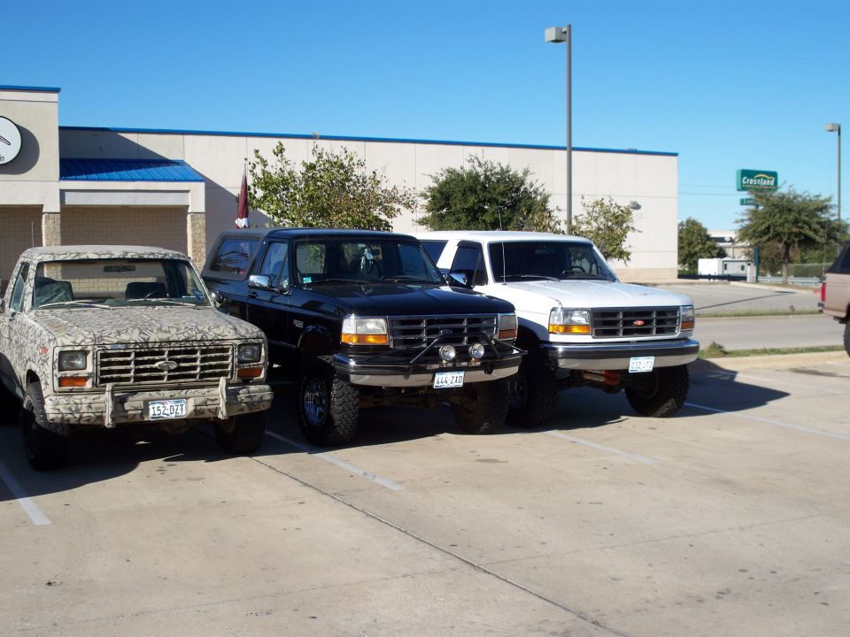 Three trucks parked in a lot. One is camo, one is black, and one is white. The white one looks like a 1996 Ford Bronco