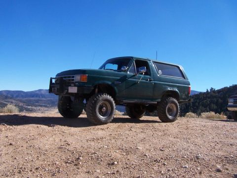 1990 Ford Bronco - A modified green Ford Bronco sits on a dusty incline under a clear blue sky, with another vehicle partially visible to the right.