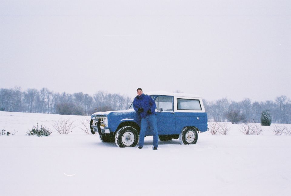 Blue and white early Ford Bronco parked in snow with a person leaning against it