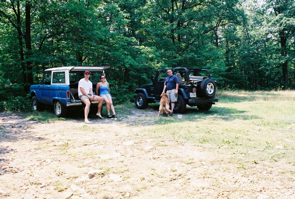 Blue early Ford Bronco and black Jeep Wrangler parked in a forest clearing with people and a dog