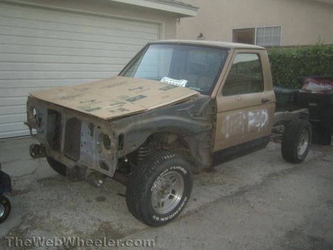 1990 Ford Bronco - tan project pickup with its front clip stripped and hood covered in cardboard sits on big tires in a residential driveway.