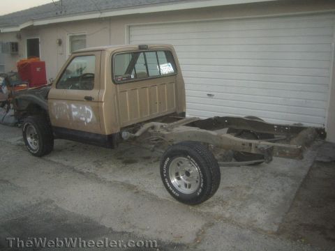 1990 Ford Bronco - tan compact pickup truck sits in a driveway with its bed removed, exposing the bare rear frame rails and chrome wheels.