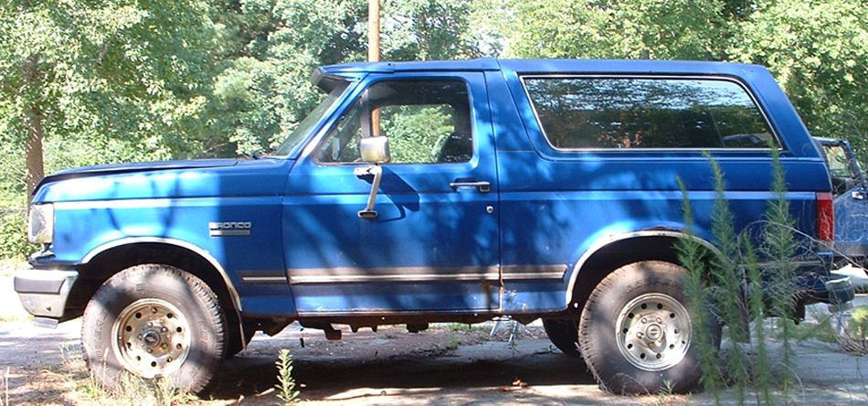 Blue Ford Bronco parked outside with trees in the background. Has a sun visor above the windshield
