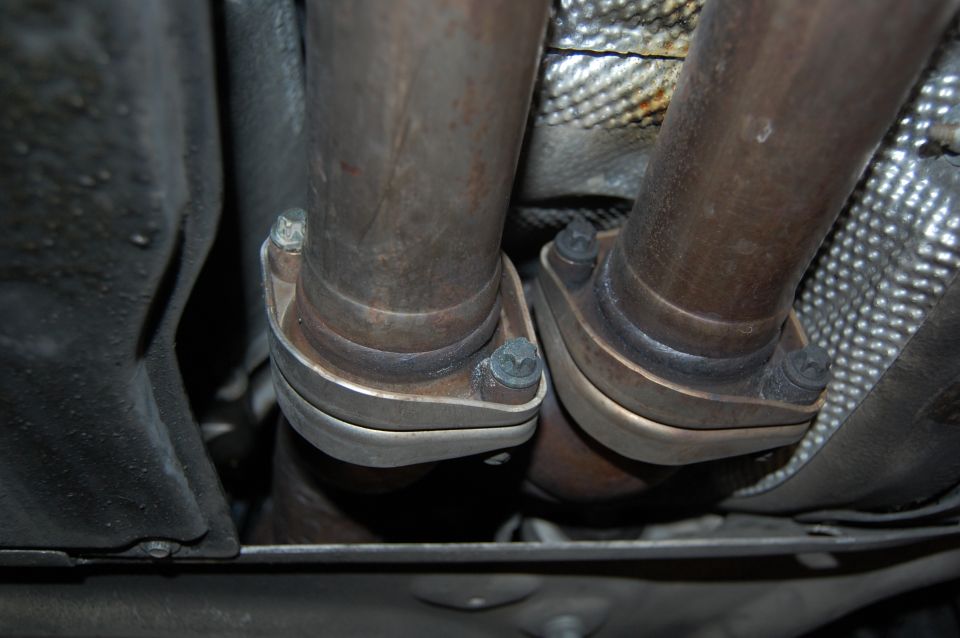 Exhaust pipe flanges and bolts on a vehicle underside, with Supermotors.net logo visible