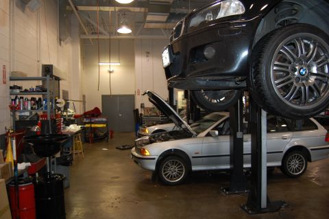 2002 BMW M3 - Two BMWs, an M3 coupe and an E39 wagon, are in a repair shop. The M3 is on a lift with its front end visible.