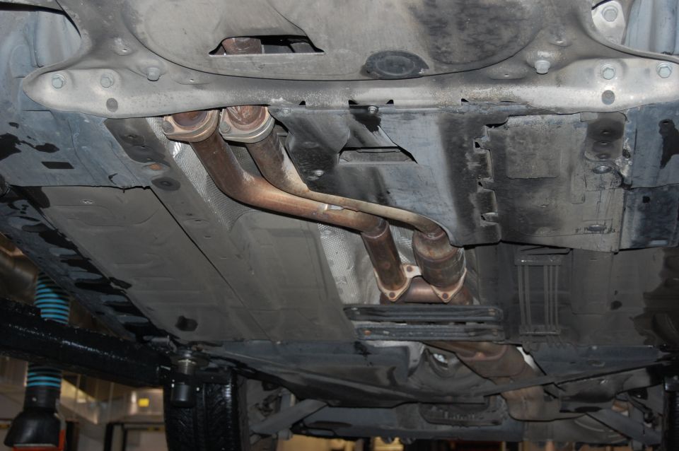 Underside of a car showing exhaust pipes and underbody panels. A Supermotors.net logo is visible