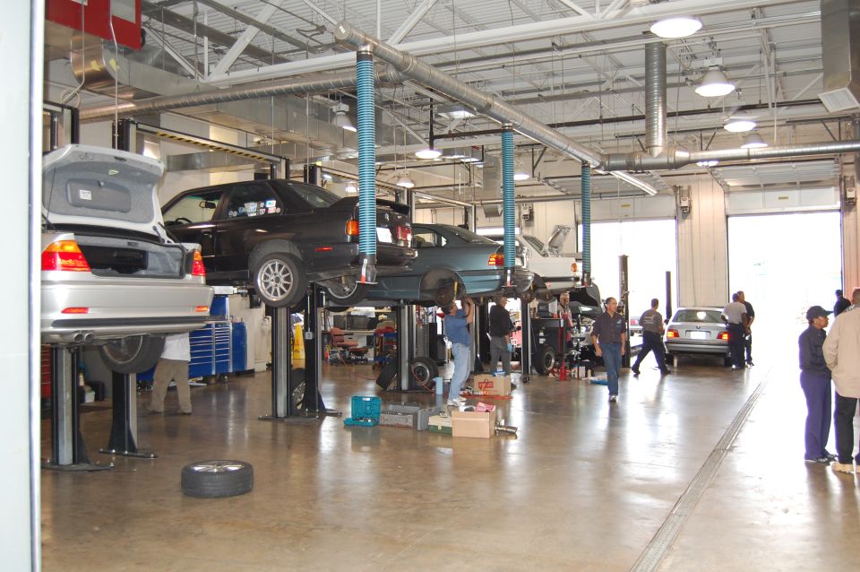 Silver BMW M3 on a lift, trunk open, in a garage with other cars and people