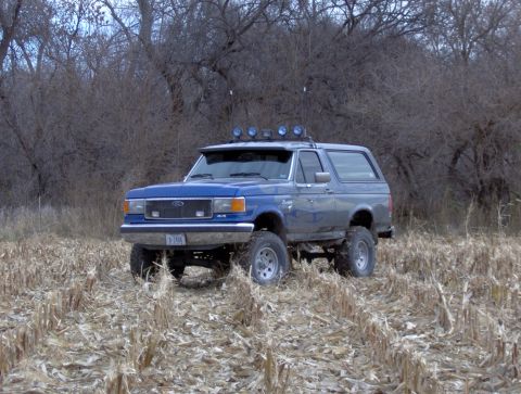 1987 Ford Bronco - This lifted blue Ford Bronco is parked in a field with bare trees in the background.