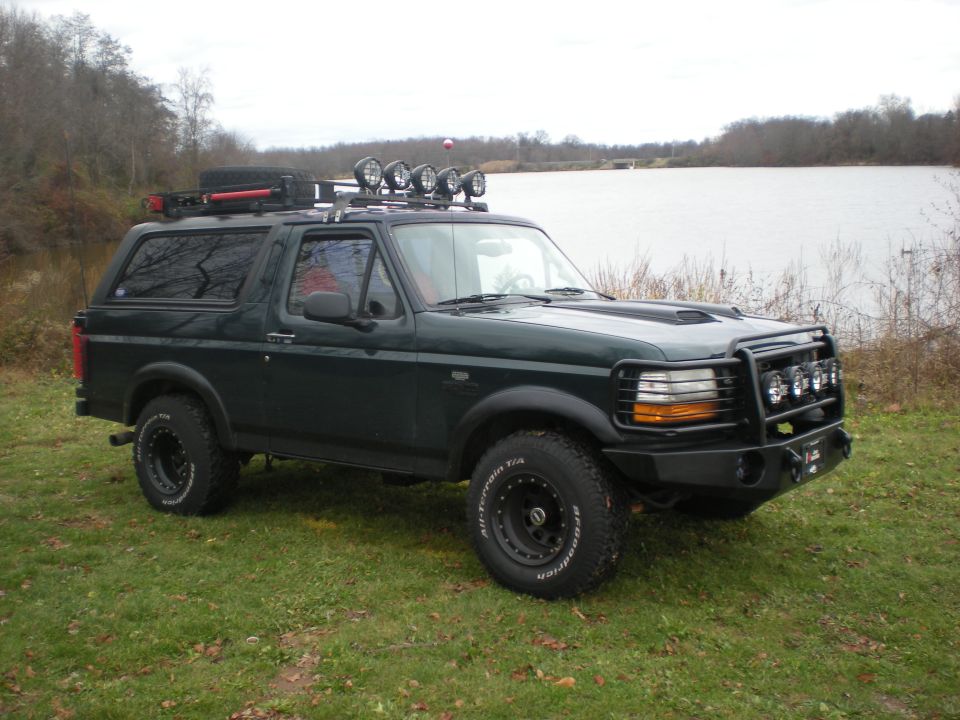 Dark green 1993 Ford Bronco with roof rack and off-road lights parked on grass near water