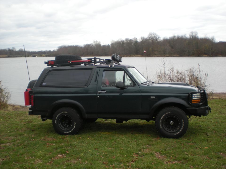 Dark green 1993 Ford Bronco with roof rack and spare tire parked on grass near a body of water