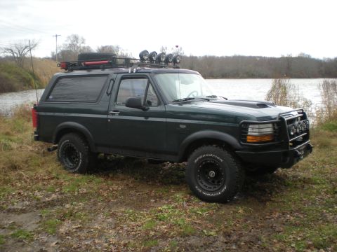 1993 Ford Bronco - This dark green 1993 Ford Bronco is equipped with a roof rack, off-road lights, and a custom front bumper, seen parked near a body of water.